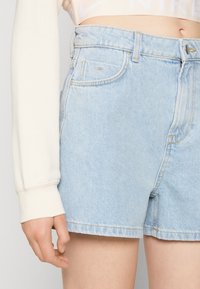 Ljusblå denimshorts med hög midja, med vanliga fickor, blekt utseende och rå nederkant. Bärs med en krämfärgad långärmad topp.