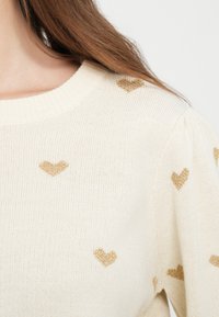 Κρεμ πουλόβερ με στρογγυλή λαιμόκοψη, διακοσμημένο με χρυσά σχέδια καρδιάς σε όλη την επιφάνεια. Απαλή υφή με φουσκωτά μανίκια και ριμπ μανσέτες.