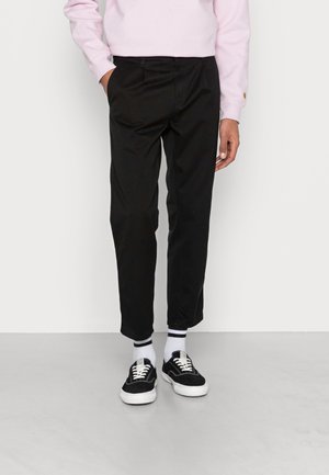 Osoba w czarnych zwężanych spodniach, białych skarpetkach z czarnymi paskami, czarno-białych sneakersach i jasnoróżowej bluzce z długim rękawem, stojąca na tle białej ściany.