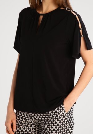 Blouse noire à manches courtes avec épaules découpées et boutons argentés, assortie à un pantalon à motifs noirs et blancs.