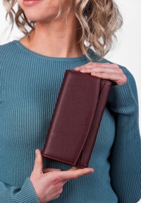 Burgundy leather wallet with a smooth texture, zip closure, and embossed logo. Held against a ribbed teal sweater.
