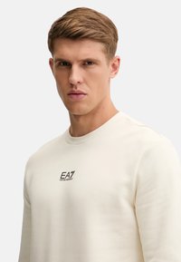 Felpa color crema realizzata in misto cotone con scollatura rotonda e maniche lunghe. Presenta un logo EA7 nero sul petto.