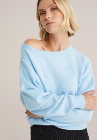 Hemelsblauwe oversized trui met een off-shoulder ontwerp, zachte textuur, geribbelde manchetten en zoom. Gedragen over een zwart gestreepte rok.
