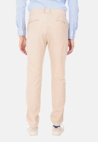Pantaloni beige con una vestibilità su misura, dotati di due tasche posteriori con chiusura a bottone, abbinati a una camicia a righe azzurre chiare.