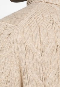 Beige stickad tröja som visar en kabelmönstertextur, med hög krage och ett mjukt, varmt utseende.