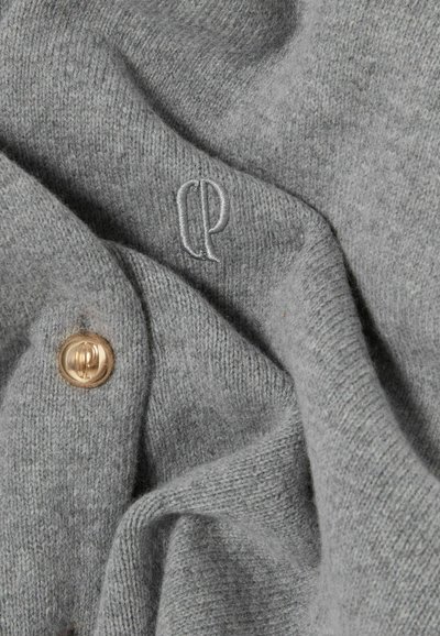 Tela de punto gris con textura suave, con un logotipo bordado "CP" y un detalle de botón dorado. Apariencia arrugada y en capas.