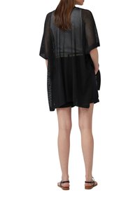 Zwarte mesh kimono-stijl cover-up met brede mouwen, losse pasvorm en open rugdetail, gecombineerd met zwarte shorts en bruin sandaaltjes.