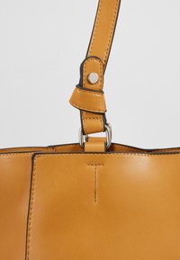 Popruh kožené kabelky v barvě opálené kůže, upevněný stříbrnými kovovými prvky a zapínáním na patent, zobrazující detailní šití a hladkou texturu.