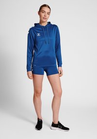 Blauer Sporthoodie mit Kordelzug, langen Ärmeln und Logo. Kombiniert mit blauen Shorts und schwarzen Sportschuhen. Glattes, leichtes Material.