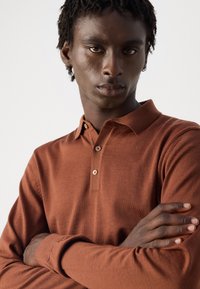 Junger Mann mit kurzen lockigen Haaren und Ohrringen, der ein rostfarbenes Langarm-Polohemd trägt und mit verschränkten Armen vor einem weißen Hintergrund steht.