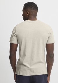 Tricou scurt cu mânecă scurtă, de culoare bej deschis, cu guler rotund și o textură netedă. Prezintă un etichete mică pe cusătura din partea stângă și un branding minim.