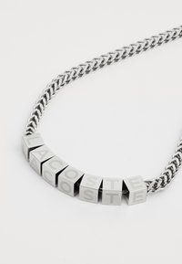 Collier en argent présentant des perles carrées avec des lettres gravées épelant "LACOSTE". Texture lisse avec des maillons entrelacés dans un design de chaîne.