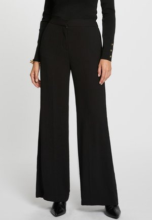 Donna che indossa pantaloni neri a gamba larga a vita alta abbinati a una maglia nera aderente a maniche lunghe e scarpe nere con punta appuntita.