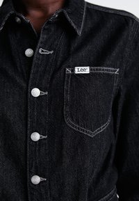 Chaqueta de mezclilla oscura con costuras blancas, botones de metal grabados con "Lee", y un bolsillo delantero que presenta una etiqueta blanca de "Lee".