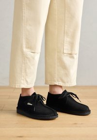 Pantofi din suede negri cu profil jos, vârf rotund, design cu șireturi și talpă texturată, asortați cu pantaloni scurți de culoare deschisă.
