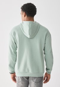 Mintgrønn hettegenser med snorhette, elastiske mansjetter og avslappet passform, kombinert med grå jeans, sett fra baksiden.