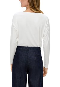 Vrouw in een lange witte blouse en donkerblauwe high-waisted broek, weergegeven van achteren tegen een witte achtergrond.