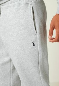 Harmaa huppari, jossa on sileä pinta; siinä on sivutaskut ja pieni musta brodeerattu logo vasemmassa yläreidessä.