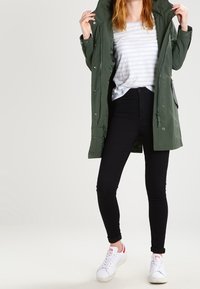 Grön parkajacka med huva över en ljus randig skjorta, kombinerad med svarta skinnyjeans och vita sneakers.