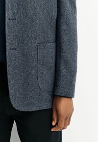 Blazer mit Fischgrätenmuster in Dunkelblau und Grau, ausgestattet mit einer Brusttasche und taillierter Passform. Das Material wirkt strukturiert und gewebt.