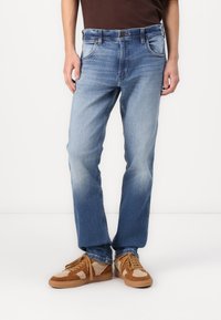 Jeans de mezclilla azul claro con corte recto, diseño desgastado, cinco bolsillos y detalles en costura marrón. Llevados con zapatillas marrón y beige.