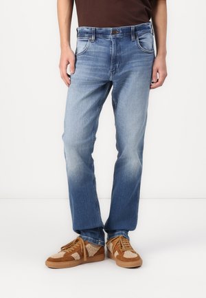 Heledsinine denim teksad sirge lõikega, kulunud disainiga, viie taskuga ja pronksist õmblusdetailidega. Kannab pruunide ja tan värvi tossudega.