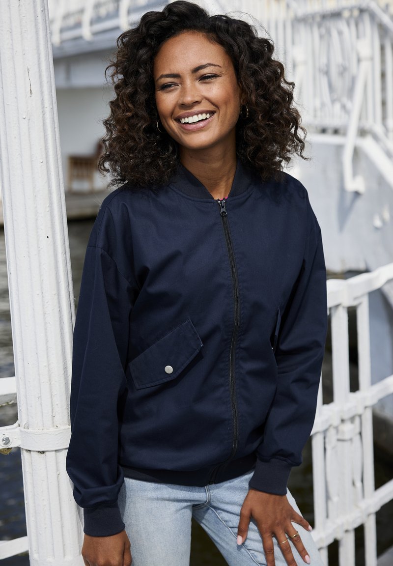 Navy bomber jacket with a zip front and a side pocket, paired with light blue jeans. The jacket features ribbed cuffs and collar.
