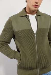 Maslinasto zeleni zip-up jakna s rebrastom teksturom na tijelu i glatkim gornjim dijelom. Ima ovratnik i bočne džepove.