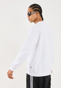 Young man with braided hair wearing black sunglasses, a white long-sleeve shirt, and black pants with white stripes, turned slightly sideways.