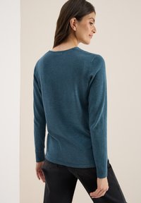 Langarm trui met ronde hals in teal, gemaakt van zacht materiaal. Heeft een relaxte pasvorm en geribbelde manchetten. G gedragen met donkere jeans.