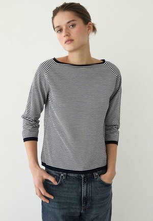 Giovane donna con capelli legati indietro che indossa una maglia a maniche lunghe a strisce bianche e nere e jeans blu scuro, in piedi con le mani parzialmente nelle tasche.