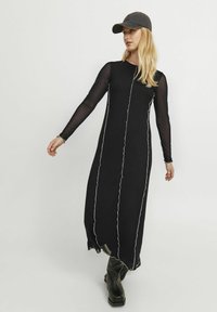 Lang zwart jurkje met lange doorzichtige mouwen, met witte stiksels langs de voorkant. Draagt een grijze pet en zwarte kniehoge laarzen.