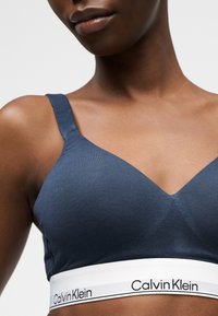 Detailný záber na tmavú osobu nosiacu námornícky modrý bralette Calvin Klein s bielym elastickým pásom a čiernym textom s logom.