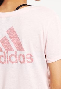 Osoba v bledě růžovém tričku Adidas s viditelným logem na zádech a černým popruhem přes rameno.