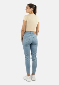 Beige eng anliegendes T-Shirt mit kurzen Ärmeln, kombiniert mit hellblauen hoch geschnittenen Jeans. Weiße Sneakers. Die Jeans hat Gesäßtaschen und ein Etikett.