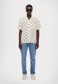 Korte mouwen, crèmekleurig overhemd met blauwe oogpatroon, gecombineerd met lichtblauwe spijkerbroek en beige sneakers. Accessoires zijn onder andere zwarte zonnebril.