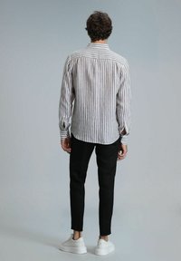 Chemise à rayures à manches longues en beige clair et marron, associée à un pantalon noir ajusté et des baskets blanches, mettant en avant une coupe décontractée et des détails au niveau des poignets.
