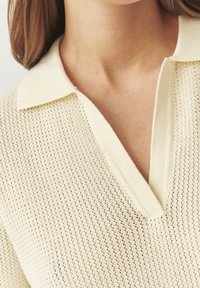 Maglione lavorato a maglia di colore crema con colletto a coste, caratterizzato da un motivo a trama aperta e un design a scollo a V. Nessun accessorio o decorazione particolare.