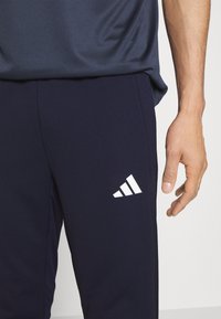 Pantalones deportivos navy con logo blanco de Adidas, hechos de un tejido suave. Cuenta con un corte relajado y bolsillos laterales. Panel de tejido texturizado en los lados.