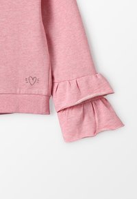 Ljusrosa sweatshirt med volangärmar och en liten broderad hjärtdekoration. Mjuk tyg med en texturerad yta.