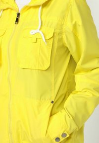 Chaqueta cortavientos amarilla con cierre de cremallera, bolsillos frontales y capucha con cordón ajustable. Presenta detalles de botones en los puños de las mangas. Textura suave.