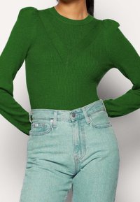 Zielony sweter prążkowany z wyraźnie zarysowanymi ramionami, w serek, połączony z jasnoniebieskimi jeansami z wysokim stanem. Gładka tekstura, dopasowany krój.