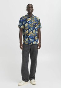 Cămașă cu mâneci scurte, cu nasturi, în albastru, cu un model floral în galben și alb, asortată cu pantaloni gri închis și adidași albi.