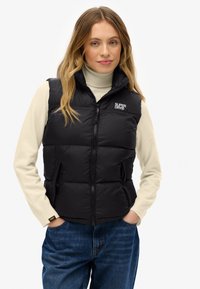 Schwarze Steppweste mit Reißverschluss, seitlichen Taschen und hohem Kragen. Getragen über einem cremefarbenen Rollkragenpullover, kombiniert mit blauen Jeans.
