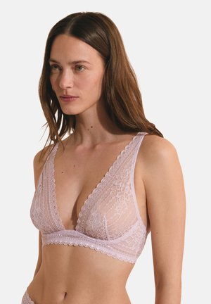 Femme aux longs cheveux bruns portant un bralette en dentelle rose clair avec des motifs floraux et des bords festonnés, debout devant un fond uni.