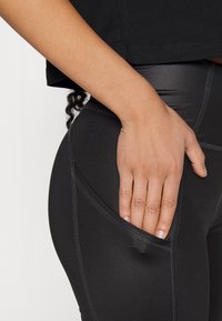 Svarta atletiska leggings med en ficka, gjorda av ett mjukt, stretchigt material och med kontrasterande sömmar. En hand vilar på fickan.