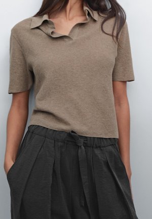 Femme portant un polo en tricot beige à manches courtes, rentré dans un pantalon noir ample taille haute avec une ceinture nouée, les mains dans les poches.
