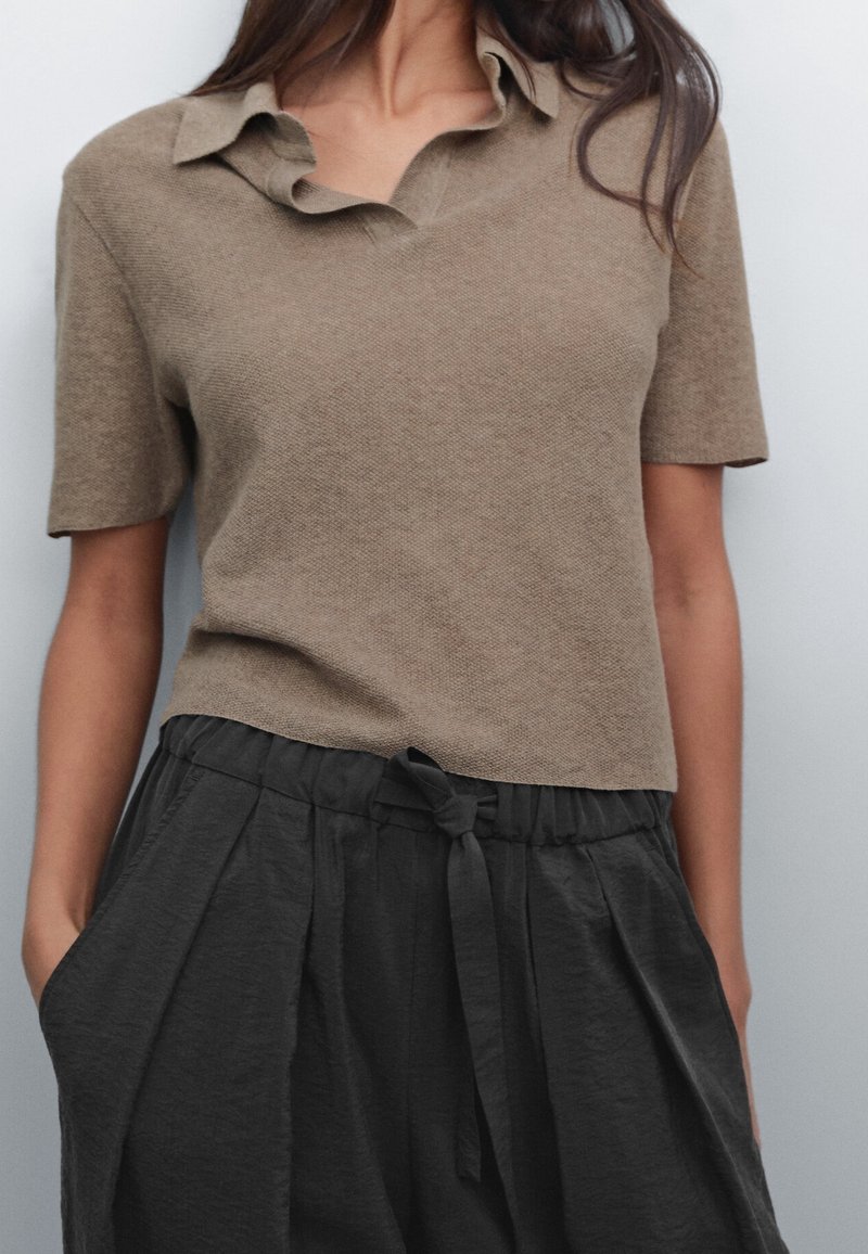 Femme portant un polo en tricot beige à manches courtes, rentré dans un pantalon noir ample taille haute avec une ceinture nouée, les mains dans les poches.