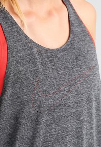 Szary sportowy top z okrągłym dekoltem, z charakterystycznym czerwonym logo Nike obrysowanym drobnymi perforacjami oraz czerwonymi akcentami na ramionach.