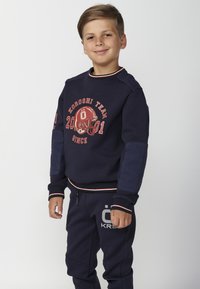 Sudadera navy con estampado gráfico rojo, cuello de canalé y parches azules texturizados en las mangas. Combinada con pantalones navy a juego que presentan un logo.